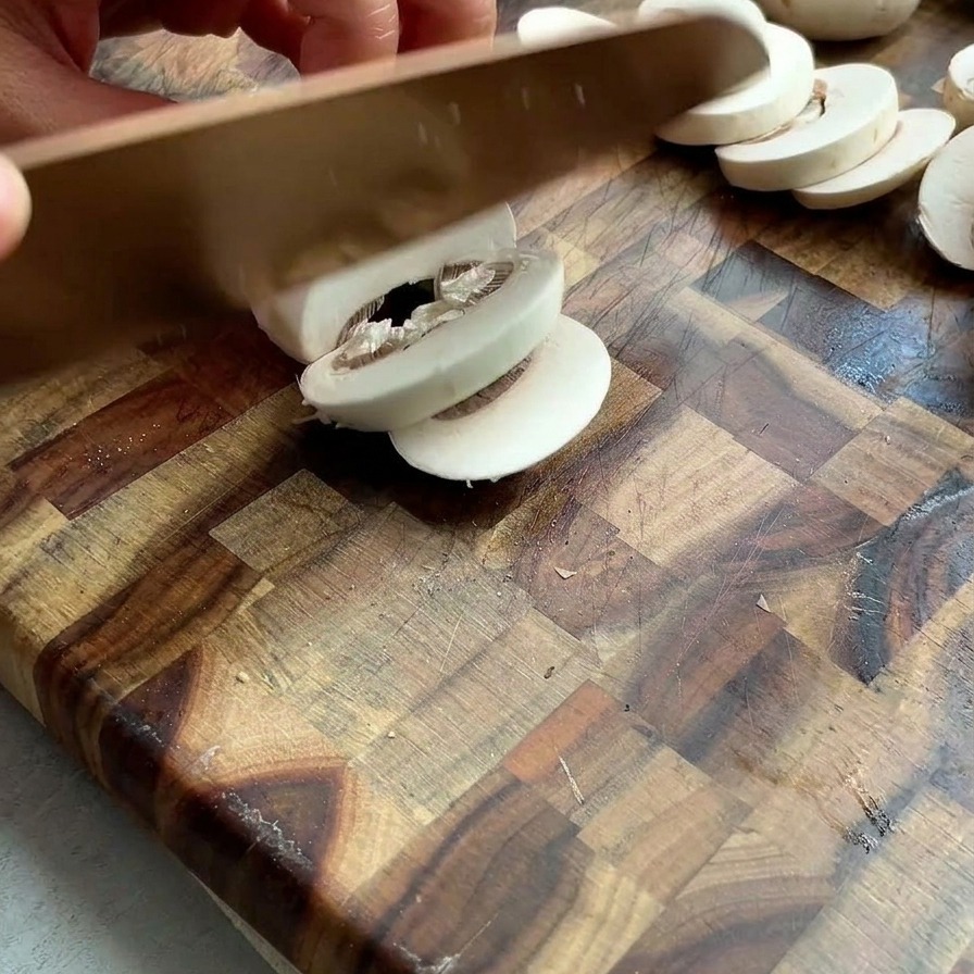 Manos cortando champiñones blancos frescos en láminas finas sobre una tabla de cortar de madera.