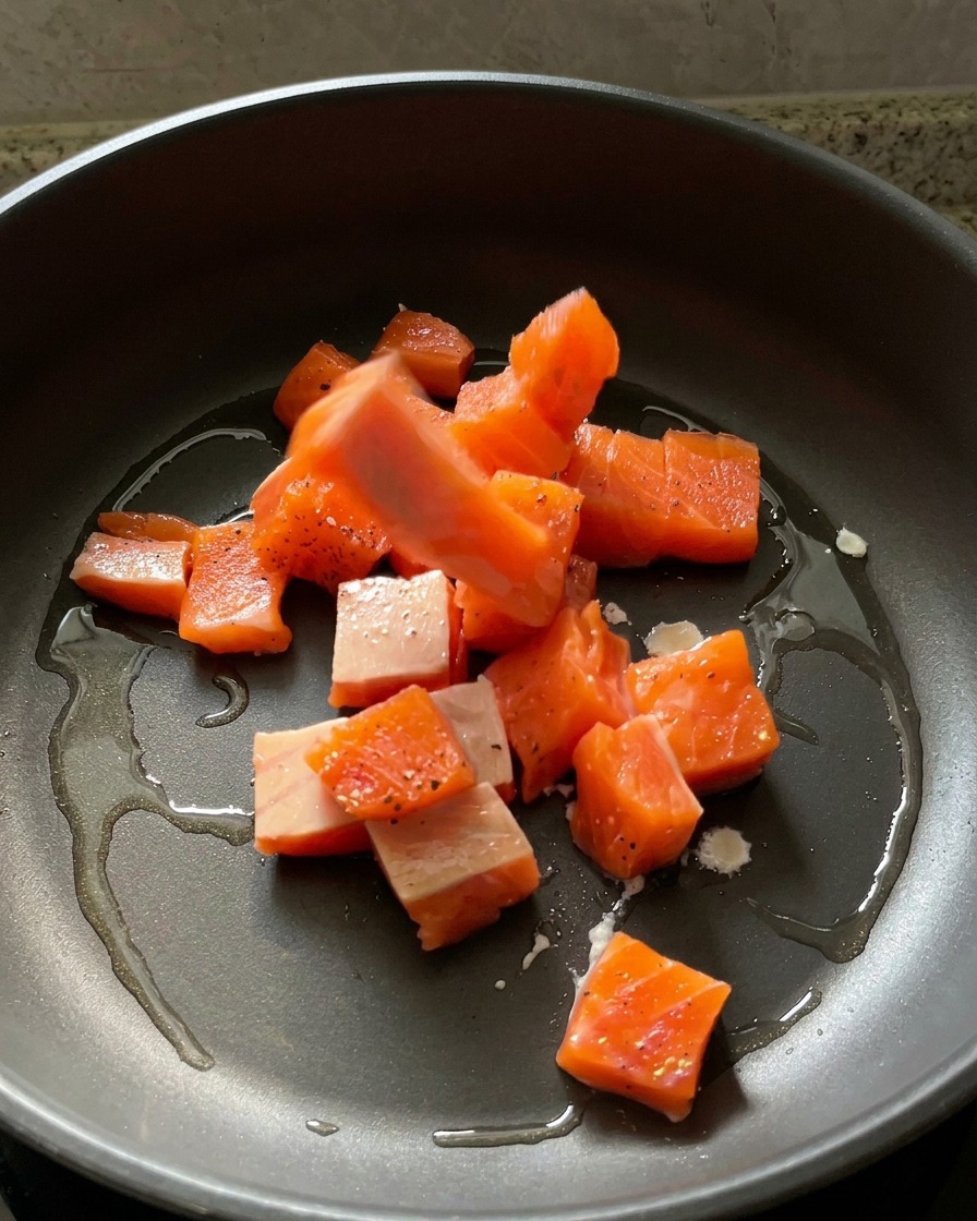 Dados de salmón crudo sazonado colocándose en una sartén oscura con aceite caliente brillante.