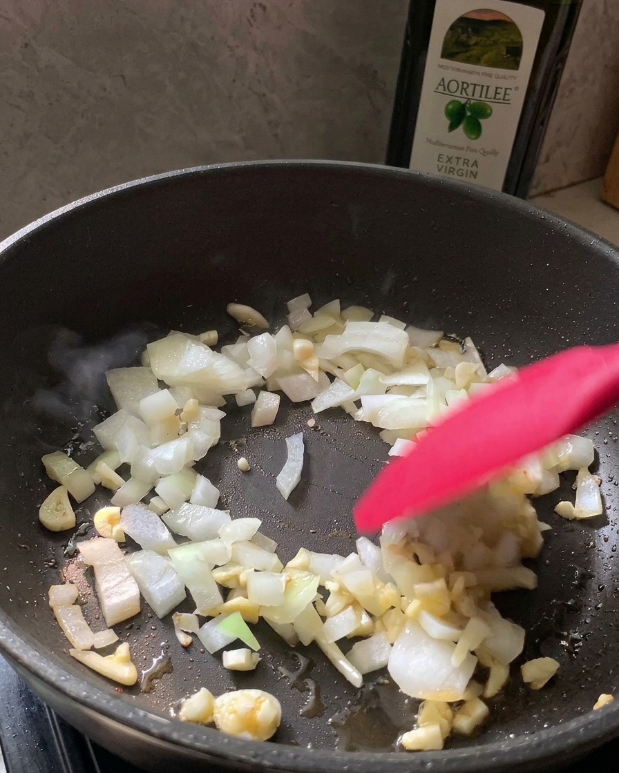 Una espátula roja removiendo cebolla blanca picada y ajo en una sartén negra.