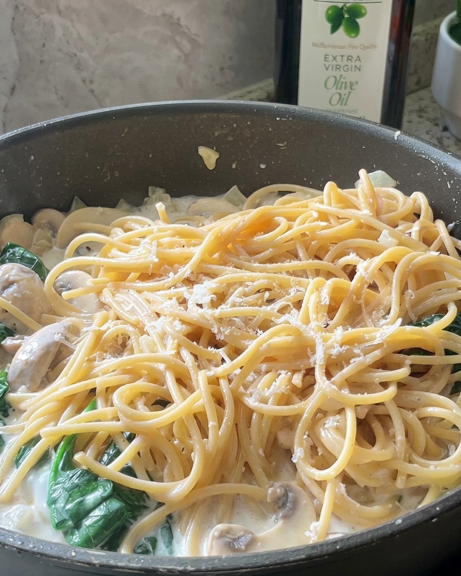 Finos hilos de queso parmesano esparcidos sobre una sartén de espaguetis cremosos con champiñones y espinacas.