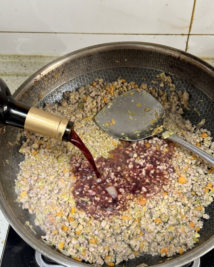 Vino tinto oscuro vertiéndose desde una botella en una sartén con carne molida dorada.