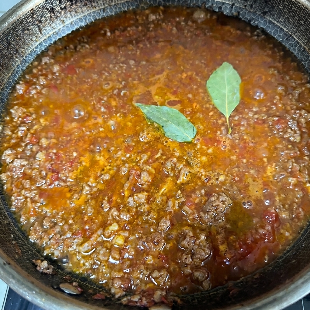 Primer plano de una rica salsa de carne burbujeando en una sartén grande con dos hojas de laurel verdes en la superficie.