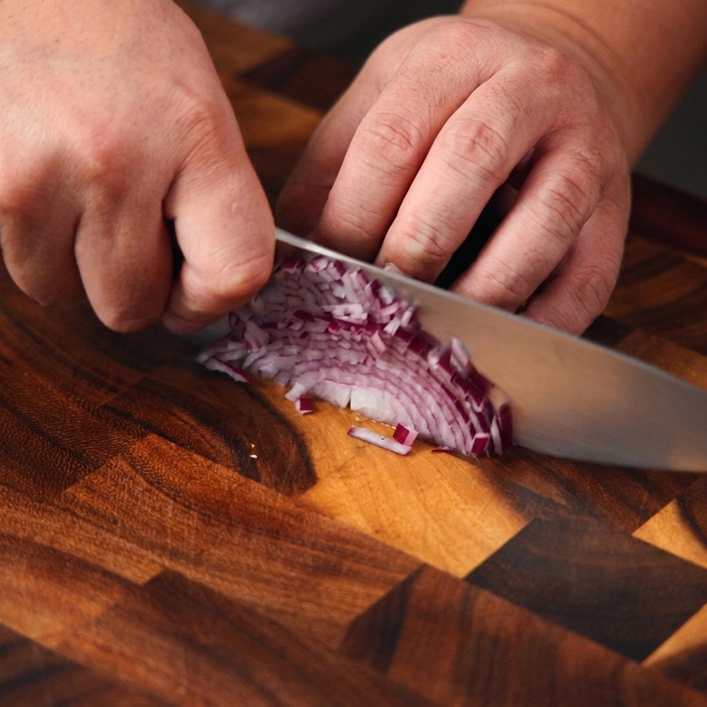Primer plano de unas manos usando un cuchillo de chef para picar finamente una cebolla morada sobre una tabla de madera.
