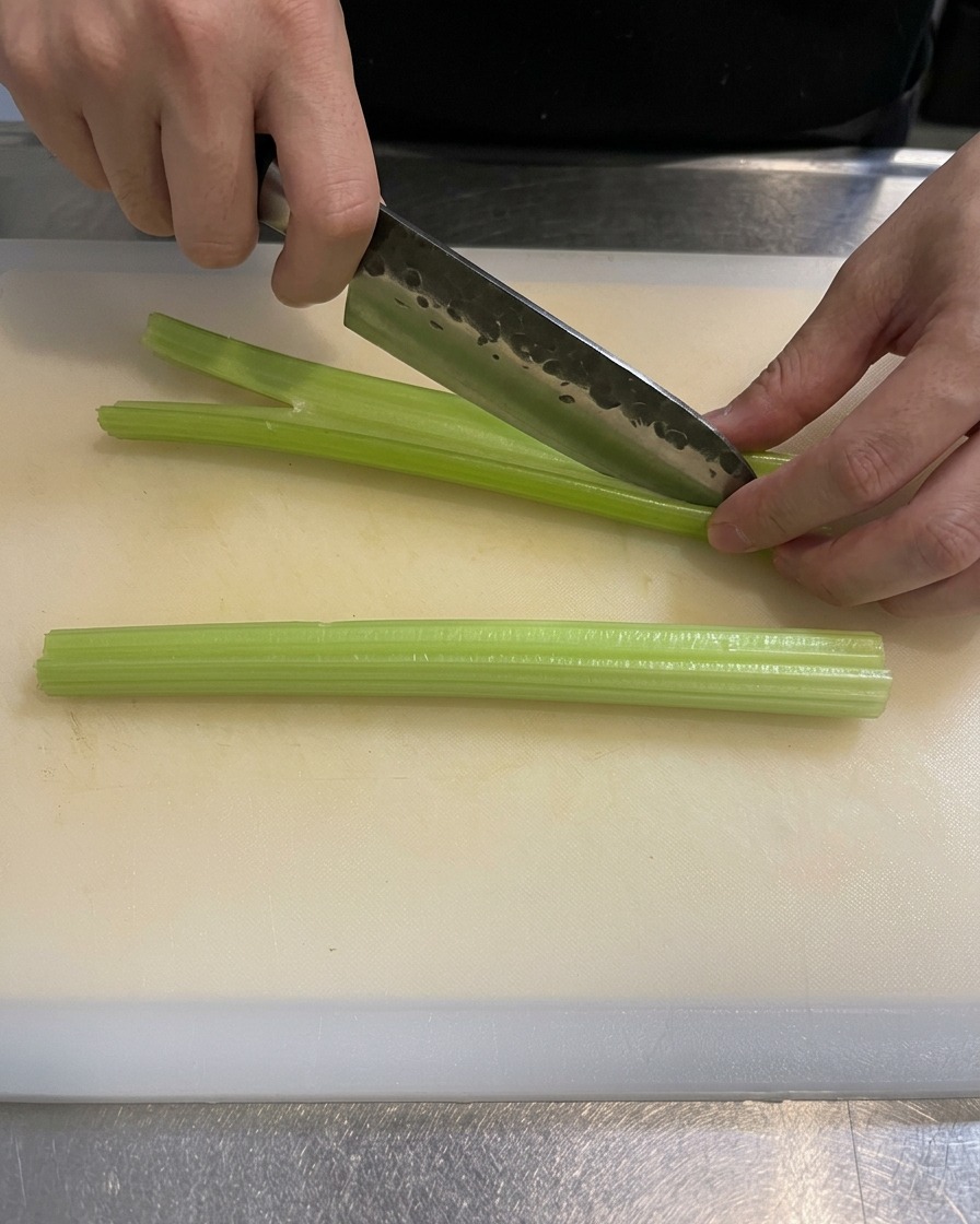 Un plano detalle de la mano de un cocinero usando un cuchillo afilado para cortar tallos de apio verde en trozos pequeños sobre una tabla de cortar blanca y limpia.