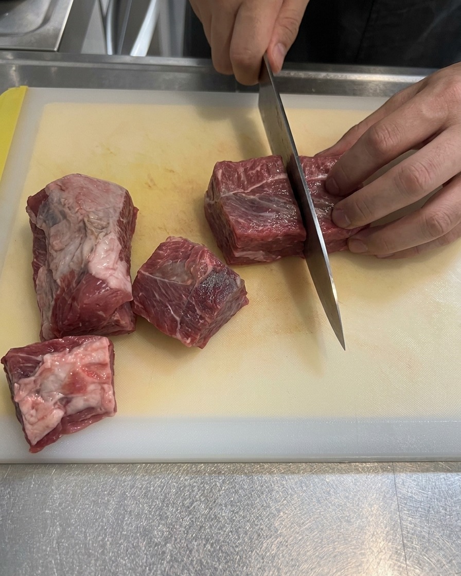 Falda de ternera cruda y veteada siendo cortada en trozos grandes cuadrados sobre una tabla de cortar blanca.
