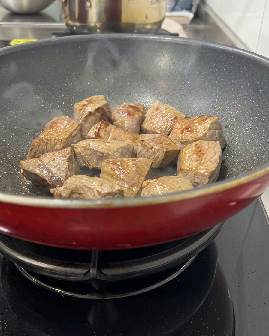 Carne de res en cubos dorándose en una sartén roja caliente, mostrando superficies doradas y un ligero vapor subiendo de la carne.