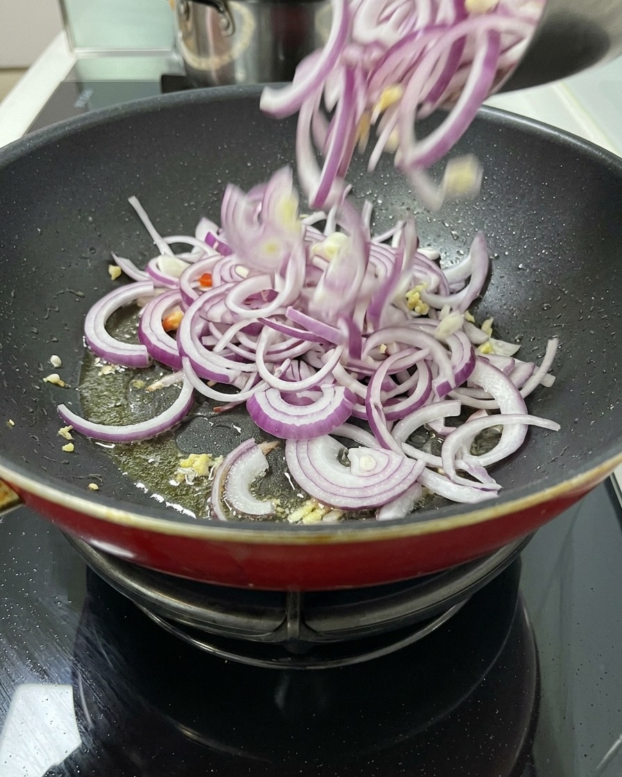 Una gran cantidad de cebolla roja cortada cayendo desde un cuenco a una sartén con aceite y vegetales aromáticos para empezar a sofreír.
