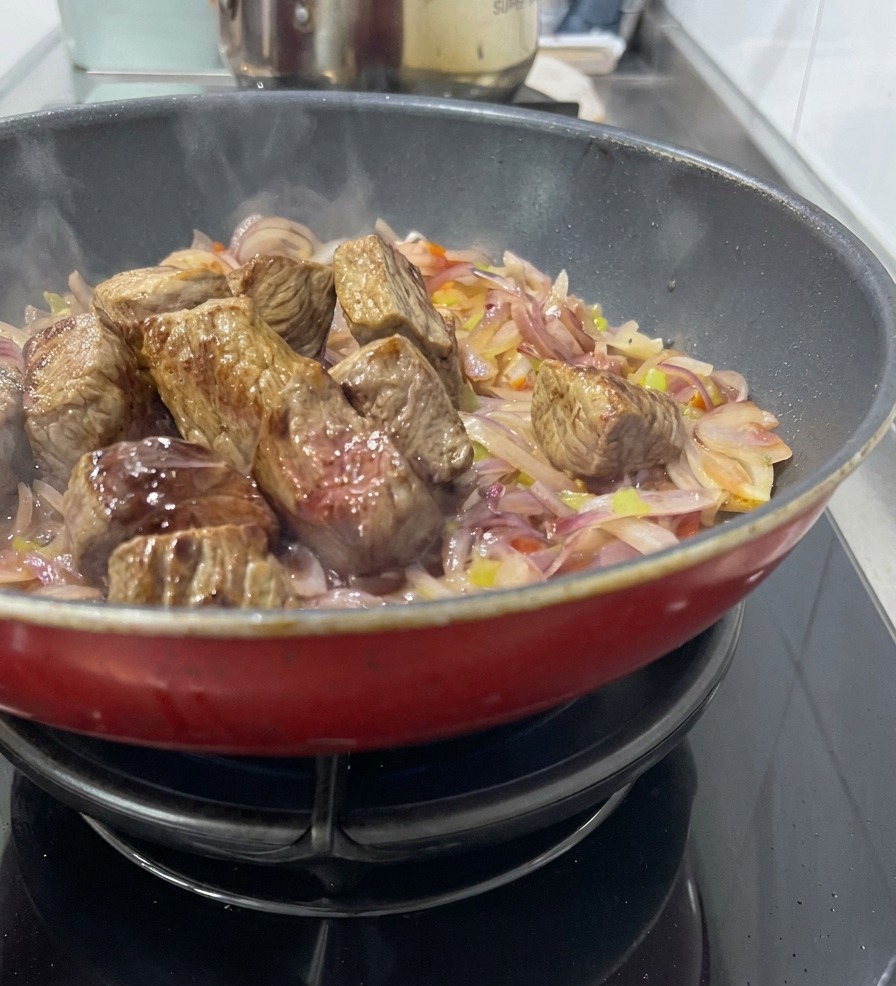 Trozos de carne sellada reposando sobre una cama de cebolla roja y apio sofritos en una sartén roja, mientras se añade vino para desglasar.