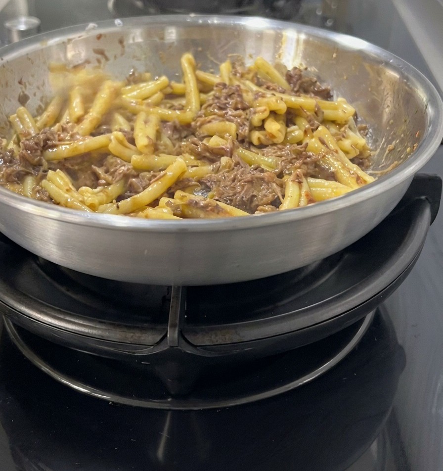 Pasta Casarecce siendo integrada en una salsa oscura y rica de carne desmechada y cebolla en una sartén de acero inoxidable.