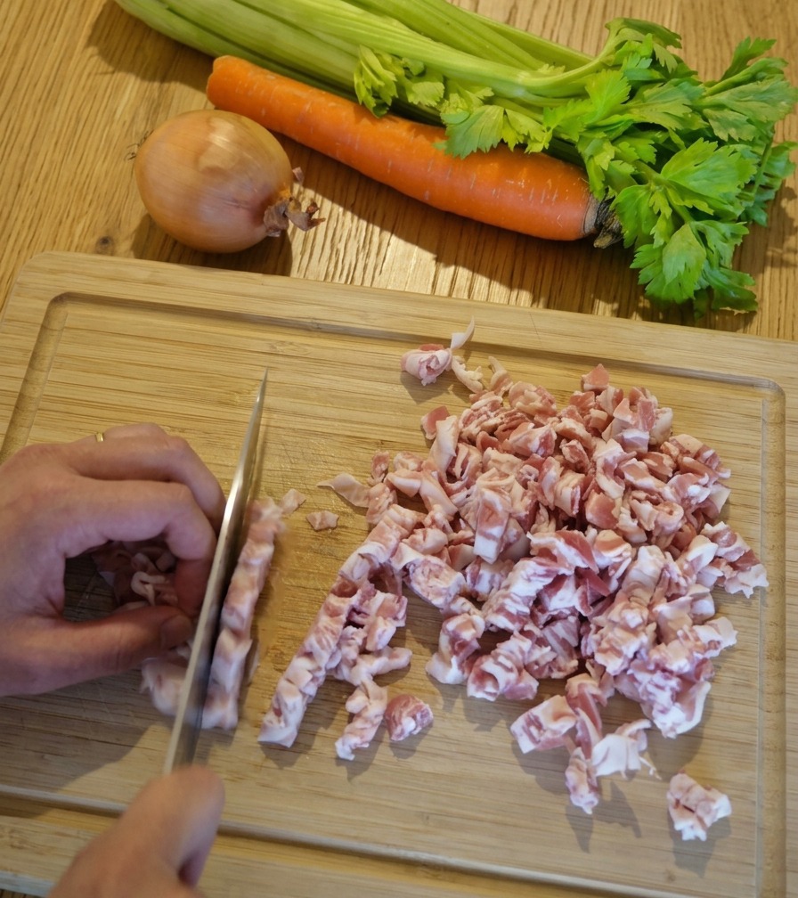 Un plano de cerca de unas manos usando un cuchillo de chef para cortar tiras de panceta rosada y blanca en dados pequeños sobre una tabla de cortar de madera clara.