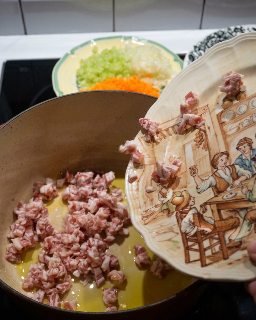 Dados de panceta cayendo desde un plato de cerámica decorado hacia una cazuela de hierro fundido roja caliente con aceite de oliva.