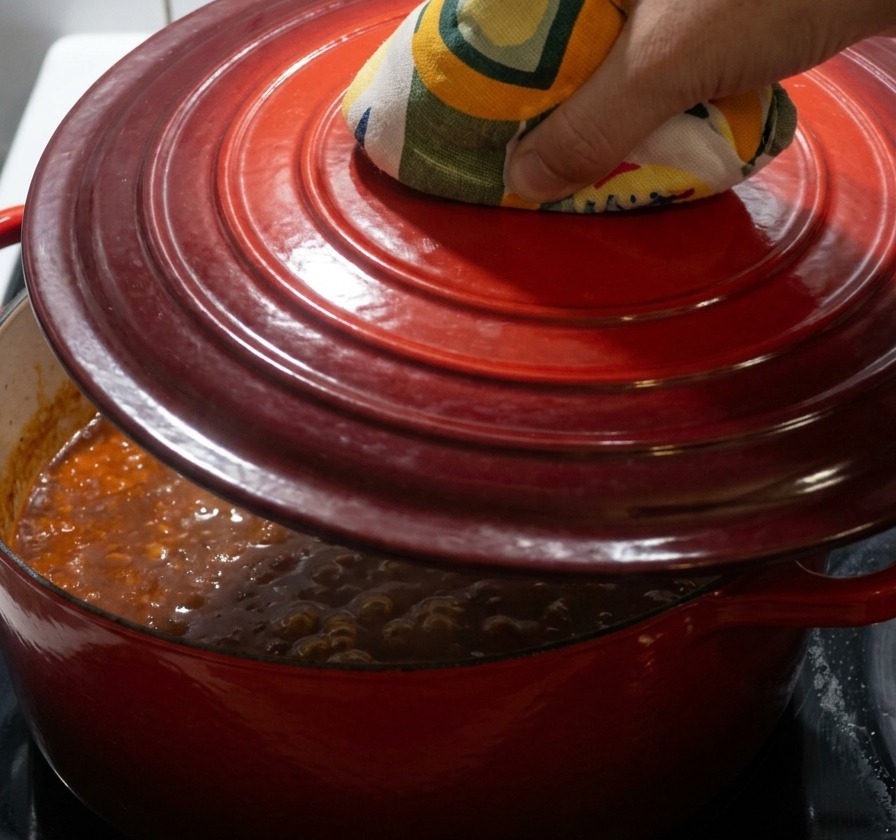 Una mano con un guante de cocina estampado en tonos amarillos y verdes colocando una tapa de hierro fundido roja sobre una cazuela que hierve a fuego lento.