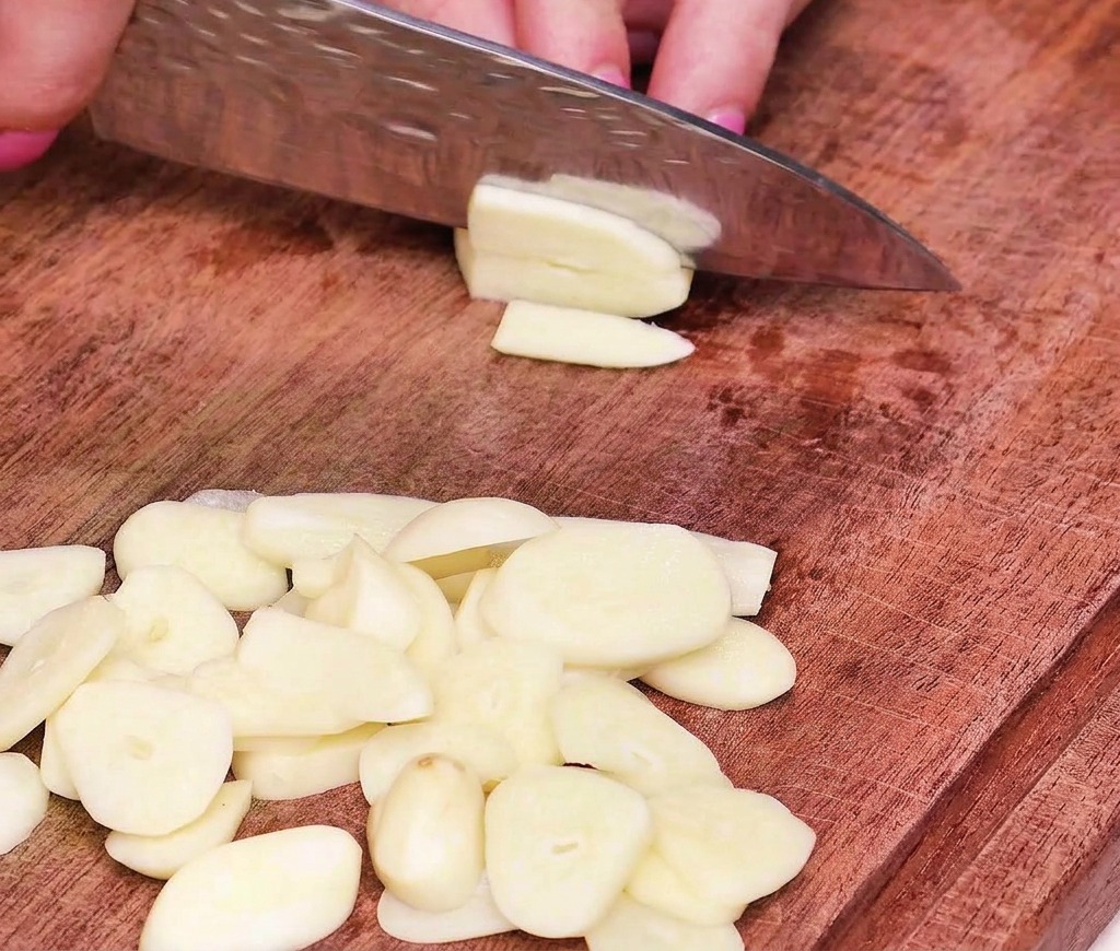 Primer plano de una mano usando un cuchillo de chef profesional para cortar dientes de ajo en láminas finas y uniformes sobre una tabla de cortar de madera.