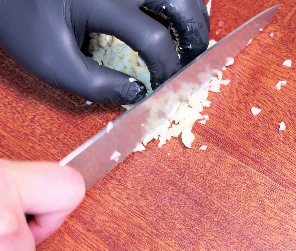 Una mano con guante negro utiliza un cuchillo de chef para picar finamente dientes de ajo crudos sobre una tabla de cortar de madera oscura.