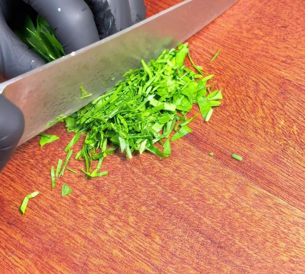 Perejil verde fresco finamente picado repartido por una tabla de cortar de madera junto al filo de un cuchillo afilado.