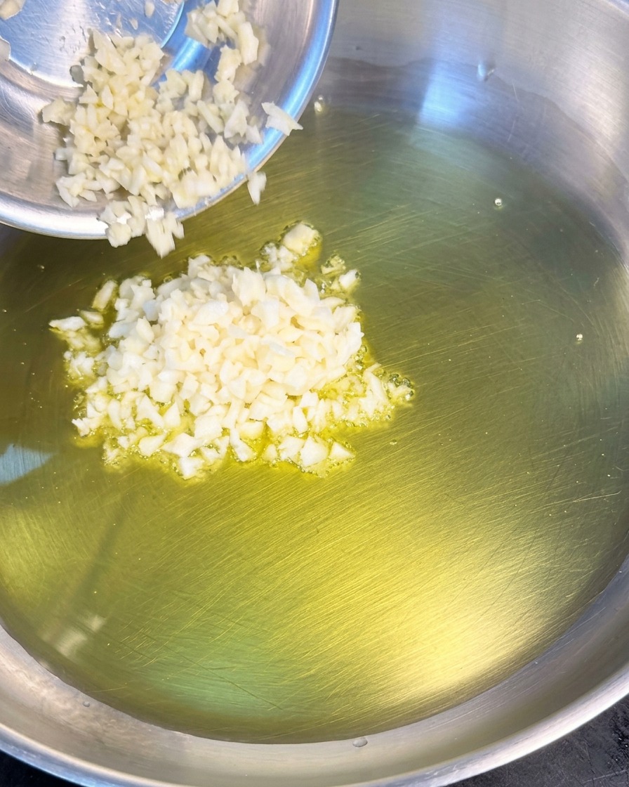 Un montón de ajo blanco finamente picado cayendo desde un pequeño cuenco metálico a una sartén con aceite de oliva brillante.