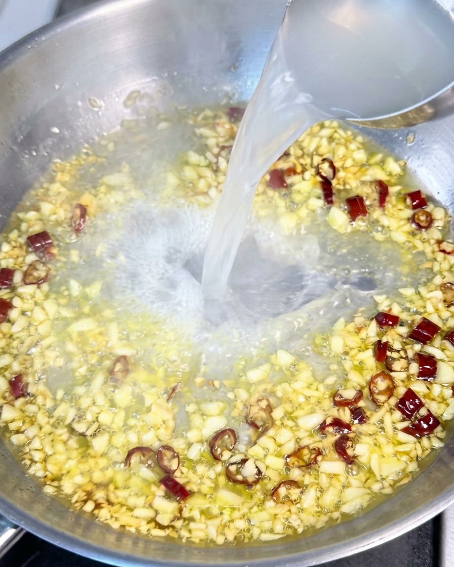Agua blanquecina de la cocción de la pasta vertiéndose desde un cucharón a una sartén con aceite, ajo y copos de chile rojo.
