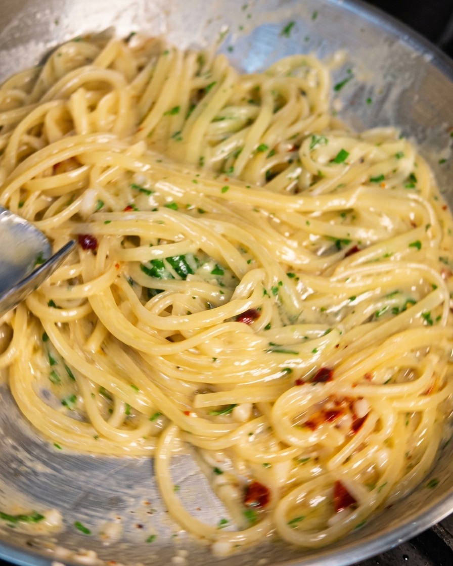 Una sartén de acero inoxidable llena de espaguetis siendo salteada con unas pinzas. Los fideos están cubiertos por una salsa cremosa y brillante con trozos visibles de ajo picado, copos de chile rojo y perejil verde fresco.