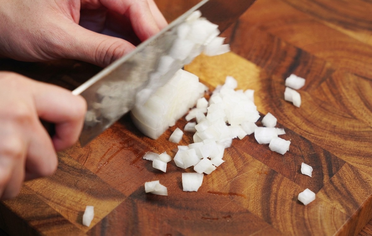 Manos de una persona usando un cuchillo de chef para picar una cebolla blanca en una tabla de cortar de madera.