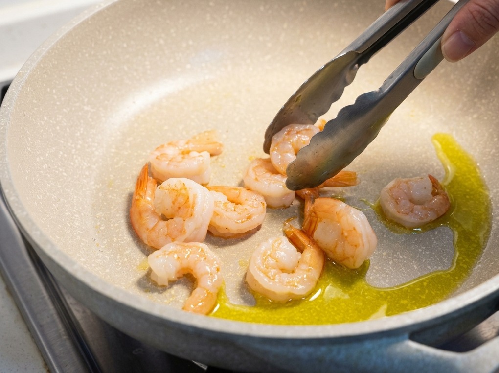 Camarones cocidos de color rosa siendo volteados con pinzas de metal en una sartén clara con aceite de oliva brillante.