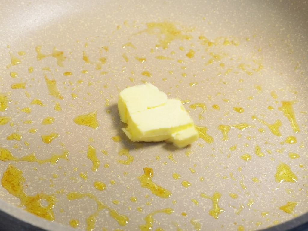 Un bloque pequeño de mantequilla amarilla derritiéndose en una sartén que contiene restos de aceite de cocina.