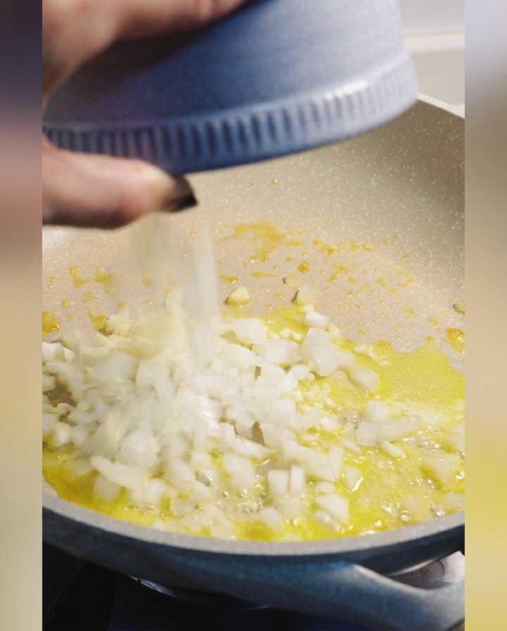 Cebolla blanca picada siendo añadida desde un tazón azul a una sartén con mantequilla derretida.