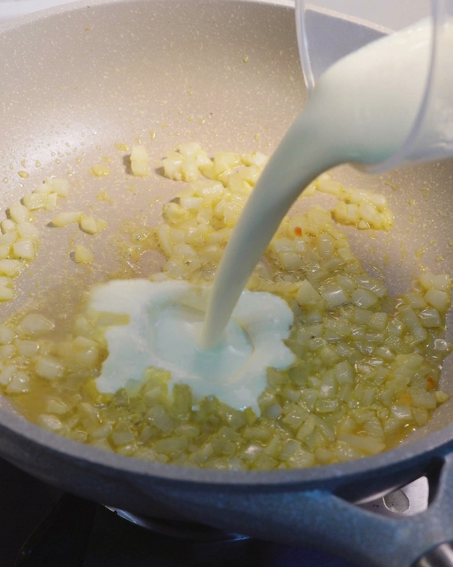 Crema de leche siendo vertida desde una taza medidora en una sartén beige con trozos de cebolla y ajo sofritos.