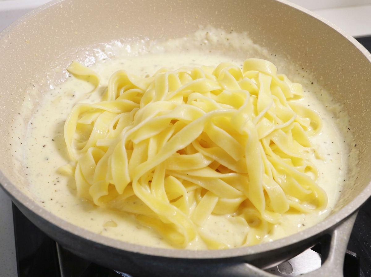 Una gran montaña de fideos fettuccine amarillos cocidos descansando sobre una salsa de crema blanca espesa y burbujeante en una sartén grande.