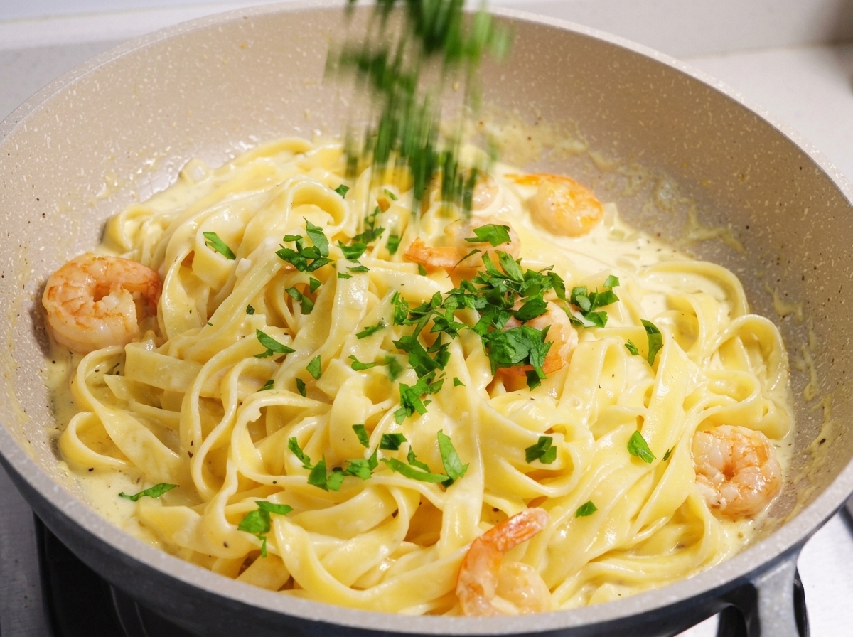 Perejil verde fresco picado siendo espolvoreado sobre un plato de pasta cremosa con camarones y fettuccine en una sartén clara.