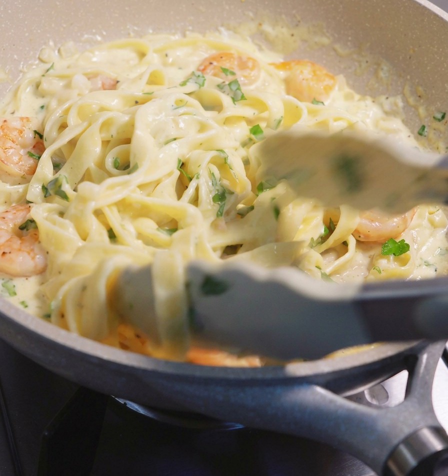 Usando pinzas para levantar y mezclar pasta fettuccine y camarones rosados en una salsa blanca cremosa dentro de una sartén moteada.