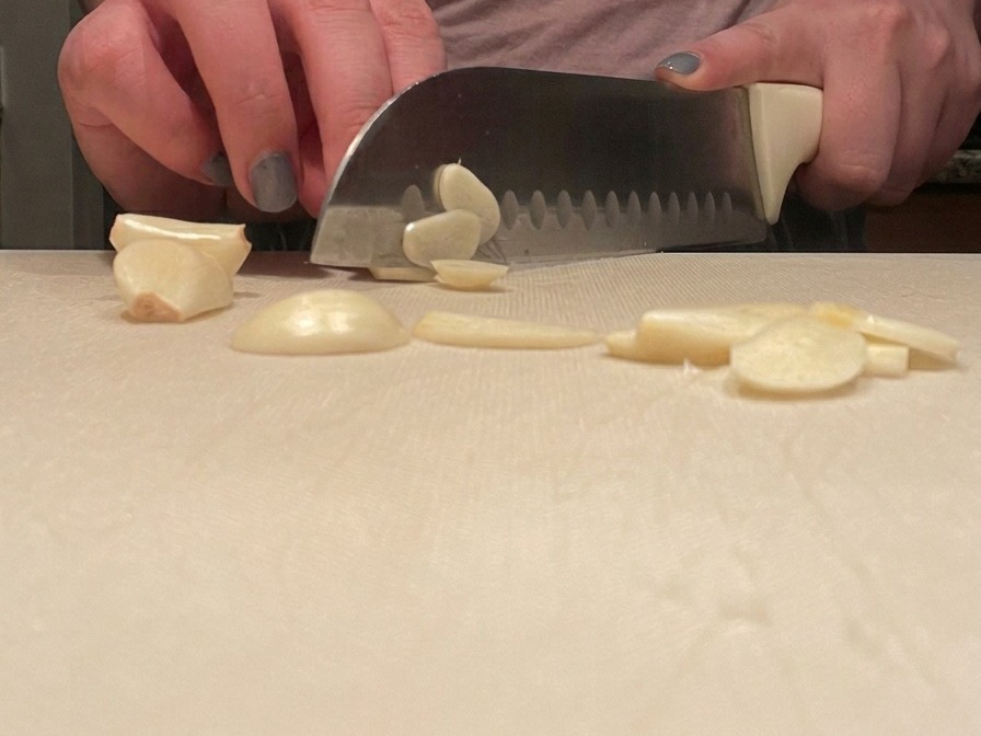 Manos de una persona cortando cuidadosamente dientes de ajo en rodajas finas con un cuchillo de chef en una tabla blanca.