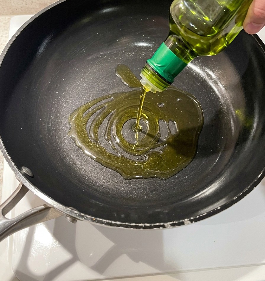 Aceite de oliva dorado vertiéndose en una sartén antiadherente oscura desde una botella de vidrio.