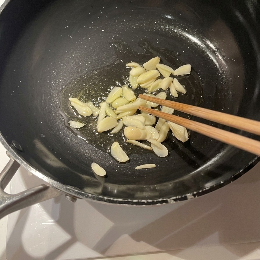 Láminas de ajo friéndose en un fondo de aceite de oliva en una sartén negra, removiéndose con palillos de madera.