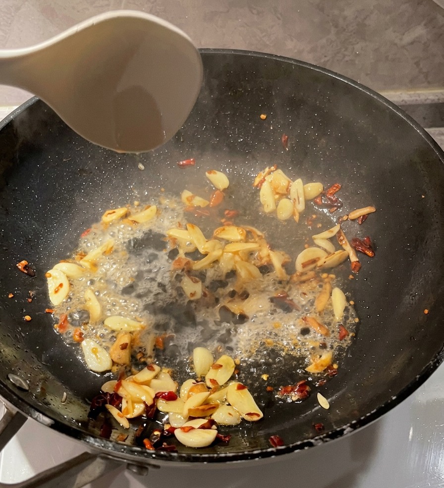 Un cucharón blanco vertiendo agua clara de la pasta en una sartén negra con láminas de ajo salteadas y copos de chile.