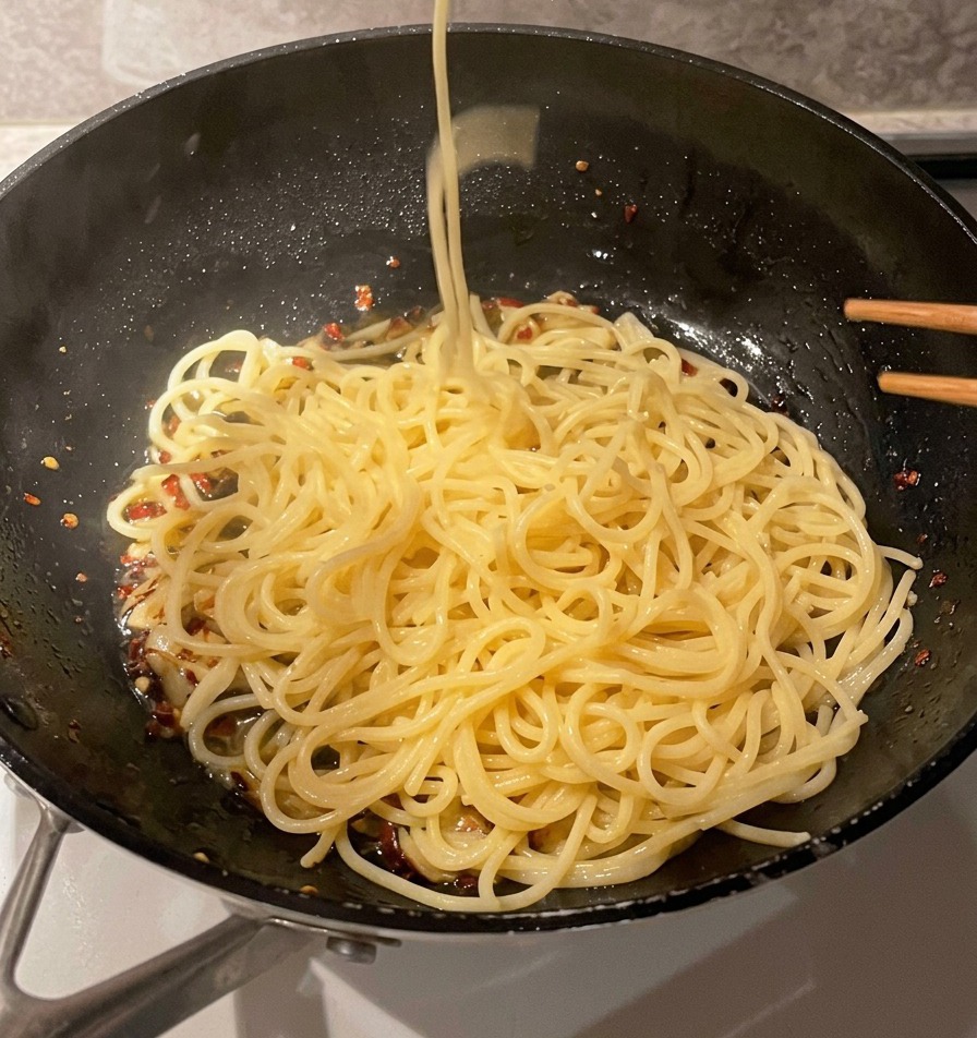 Un colador de malla metálica dejando caer espaguetis cocinados en una sartén con ajo y aceite.