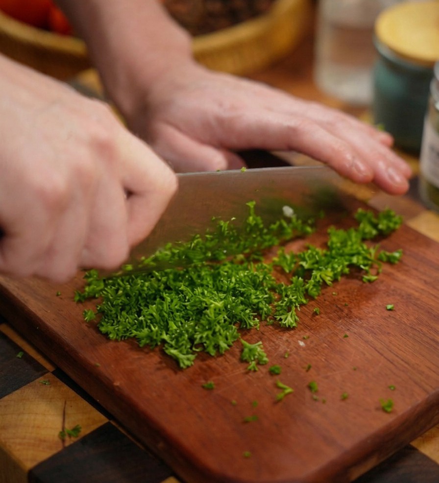 Primer plano de unas manos picando finamente perejil verde brillante con un cuchillo grande sobre una tabla de madera.