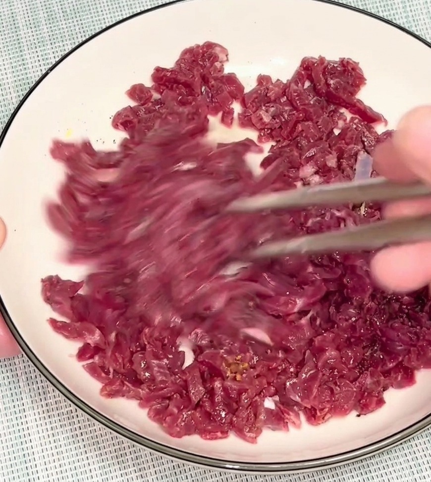 Una persona usando palillos plateados para mezclar la carne picada cruda en un bol blanco distribuyendo el marinado.