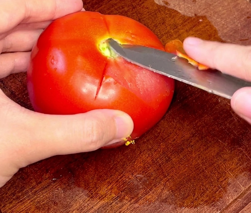 Manos retirando con cuidado la piel de un tomate entero escaldado usando un cuchillo de puntilla.