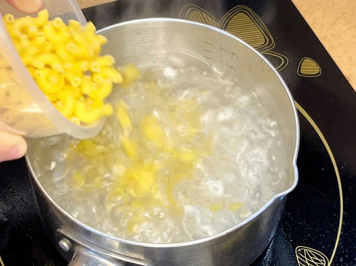 Macarrones crudos cayendo desde un recipiente en una olla de acero inoxidable con agua hirviendo.