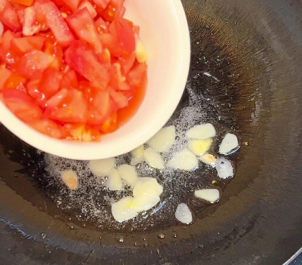 Tomates en dados vertiéndose desde un bol blanco en una sartén con aceite caliente y láminas de ajo dorado.