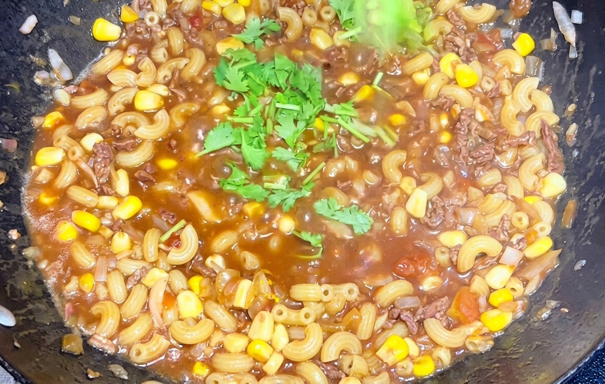 Una mano espolvoreando cilantro verde picado sobre una sartén humeante de macarrones con carne y tomate.