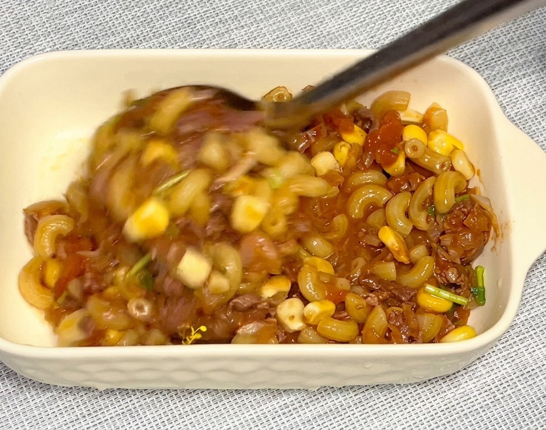 Una cuchara metálica grande trasladando la mezcla de macarrones, maíz y carne a una fuente rectangular de cerámica blanca.