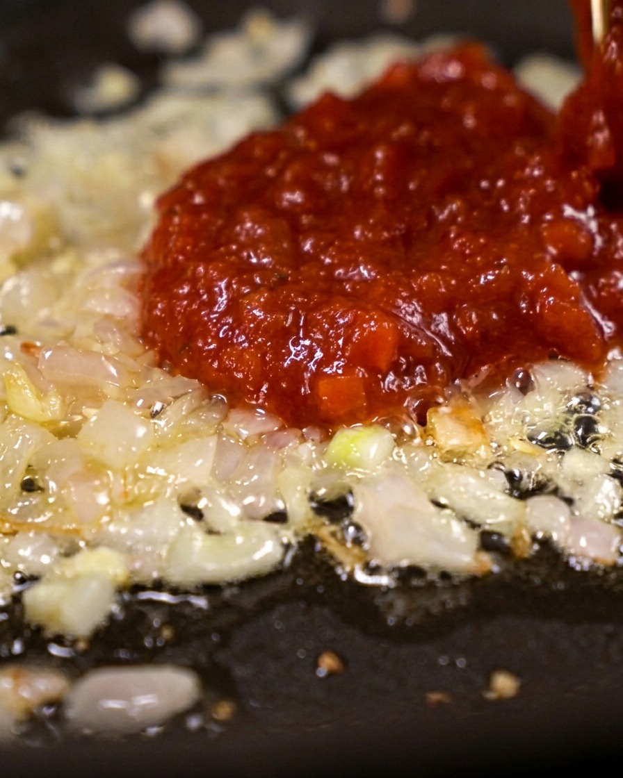 Una cucharada grande de pasta de tomate espesa y de color rojo brillante cayendo sobre un lecho de chalotas salteadas.