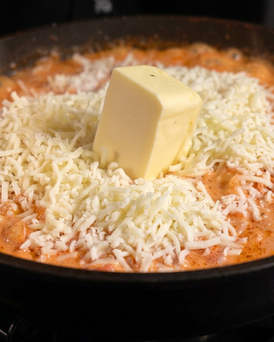 Un trozo cuadrado y grueso de mantequilla pálida colocado sobre un montón de queso mozzarella blanco rallado sobre salsa de pasta naranja.