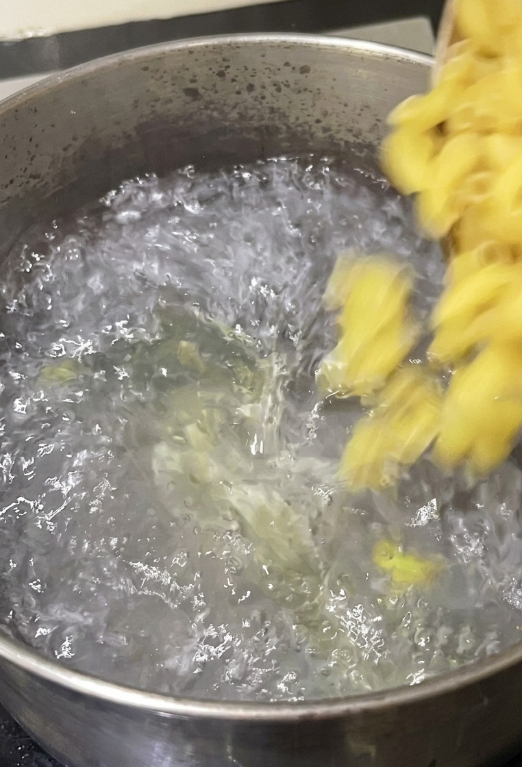 Macarrones de codito vertiéndose desde un cuenco en una olla de acero inoxidable con agua hirviendo en la estufa.
