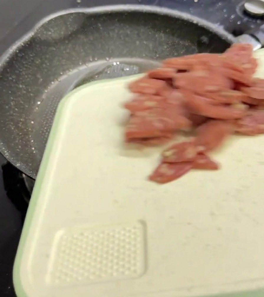 Rodajas de salami rosa cayendo desde una tabla de cortar blanca en una sartén oscura y moteada.