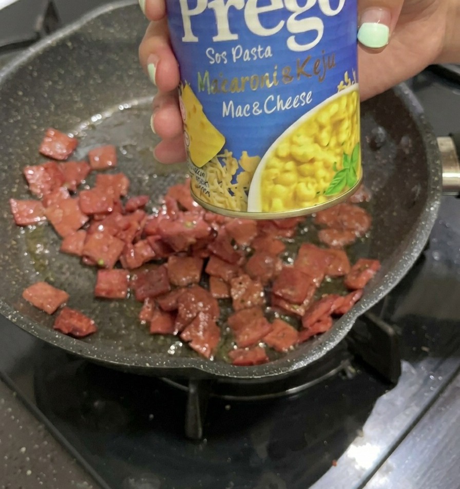 Una mano sosteniendo una lata azul y amarilla de salsa para pasta Prego Mac & Cheese sobre una sartén que contiene trozos de salami frito.
