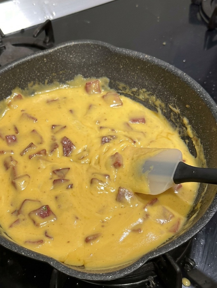 Una sartén antiadherente negra llena de macarrones de codito, salami de ternera en cubos y una salsa de queso amarilla espesa y vibrante que se remueve con una espátula de silicona blanca.