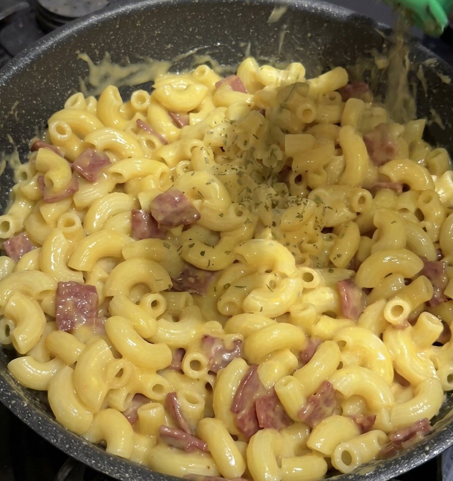 Copos de perejil verde seco cayendo desde un bote de especias verde sobre una sartén llena de macarrones con queso.