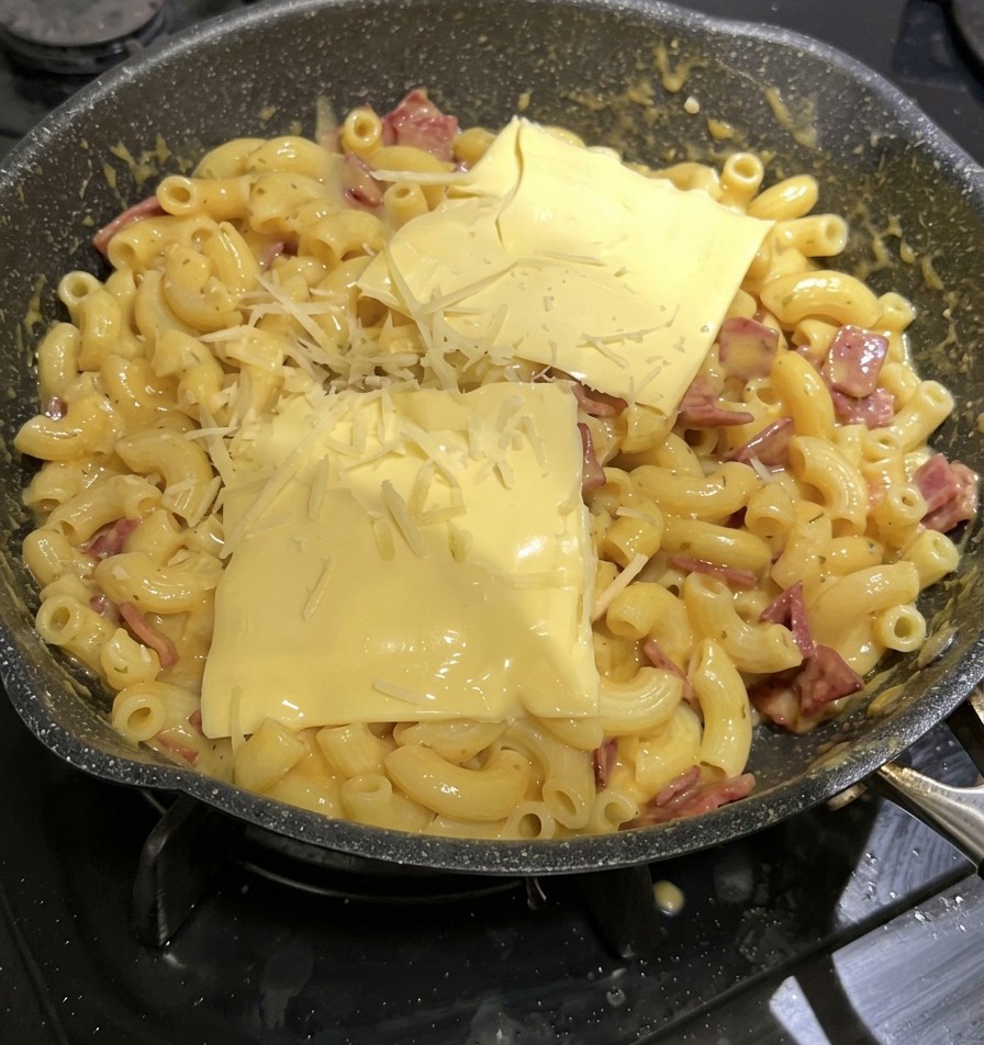 Dos lonchas amarillas cuadradas de queso procesado y queso rallado encima de una sartén de macarrones con queso.